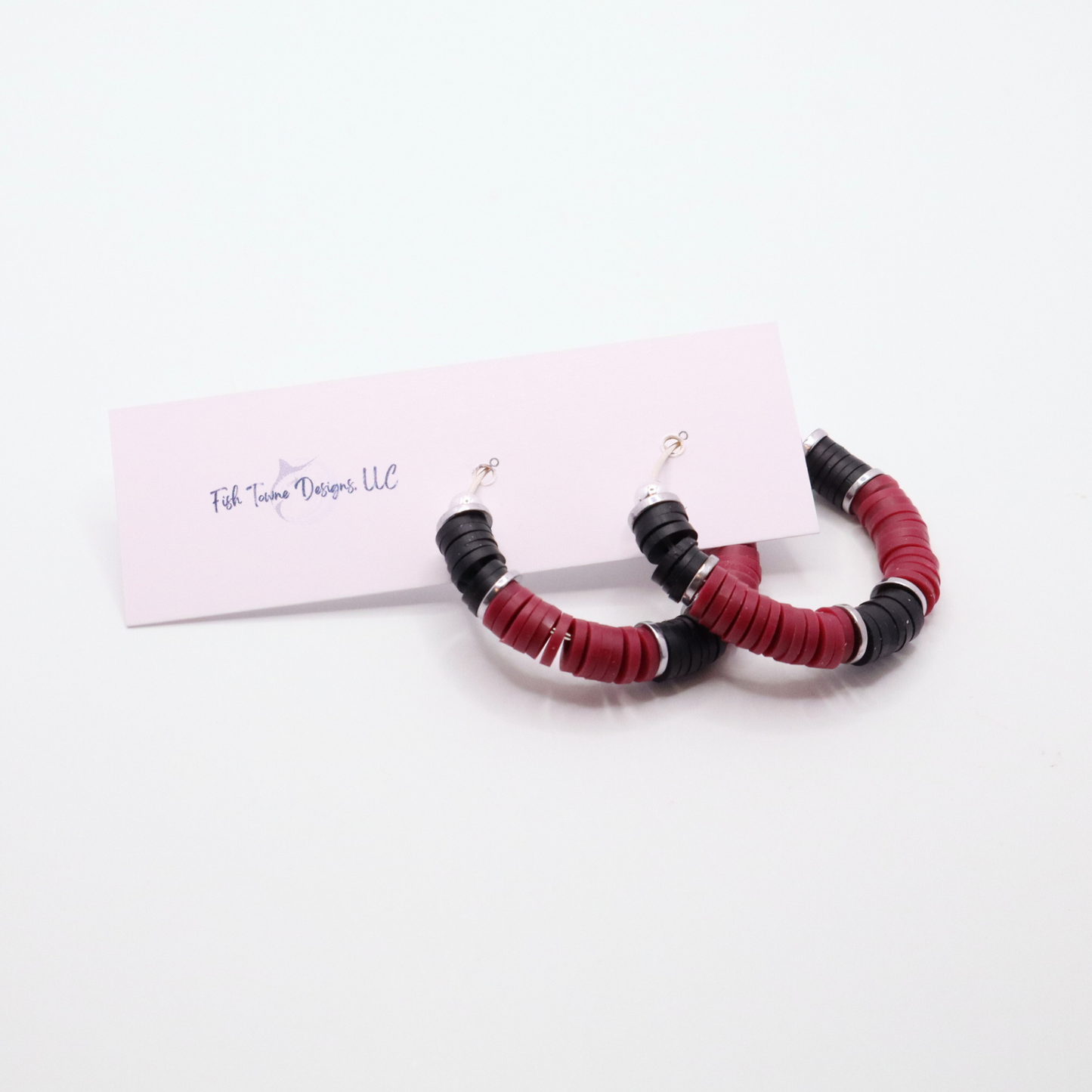 Maroon and black heishi bead hoop earrings with silver accents hanging on a minimalist jewelry stand against a white background