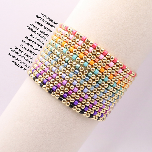 Stacked 18k gold filled beaded bracelets with colorful accent beads displayed on a wrist, labeled with color options including hot hibiscus, soft flamingo, coral blush, mango sorbet, Caribbean aqua, blue pearl, moonlit tide, Carolina sky, lilac breeze, island orchid, shoreline violet, purple plunder, and pirate flag