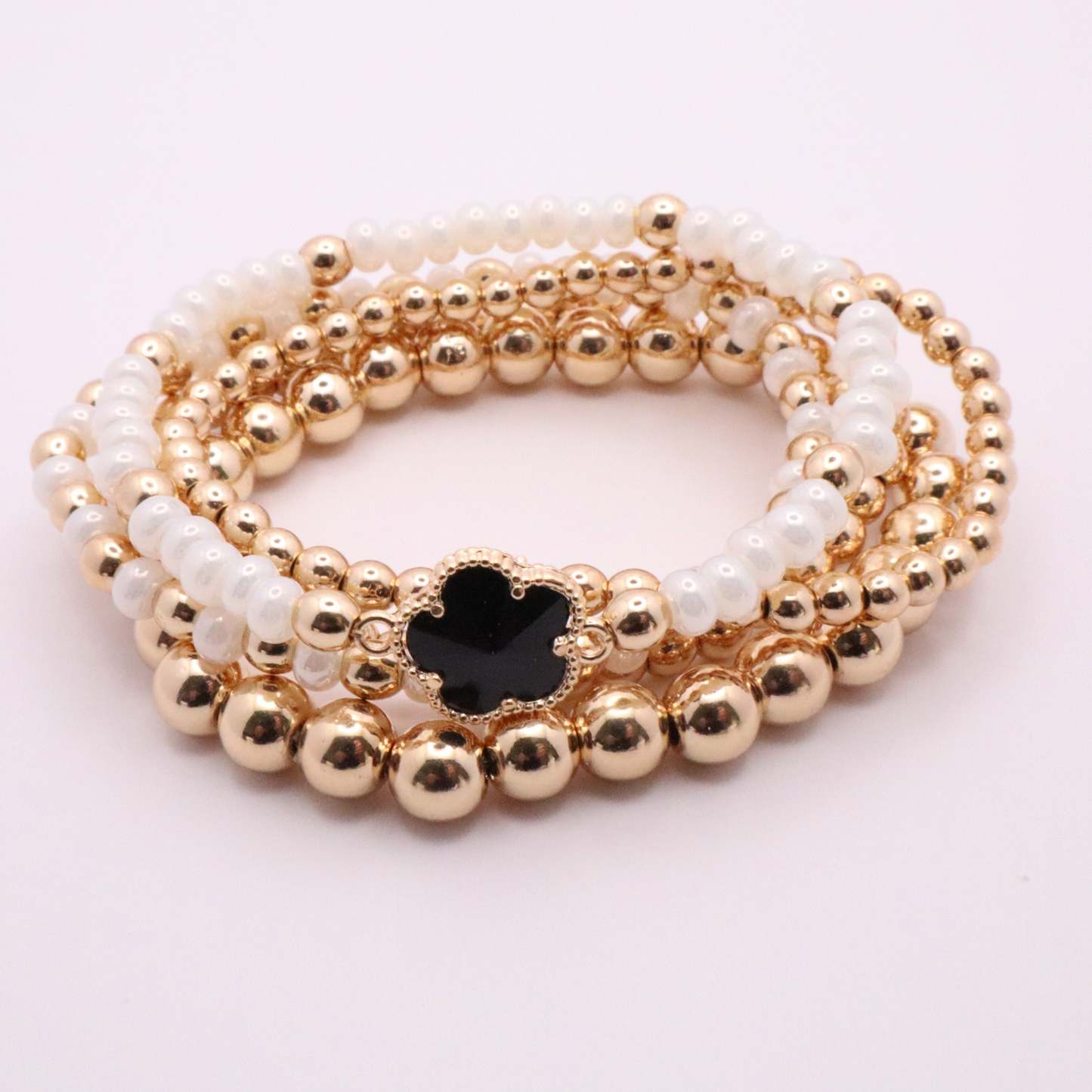 Set of 5 of gold toned beaded bracelets with white seed beads and hematite gold accents. One bracelet features a black clover charm. The bracelets are displayed on a white background.