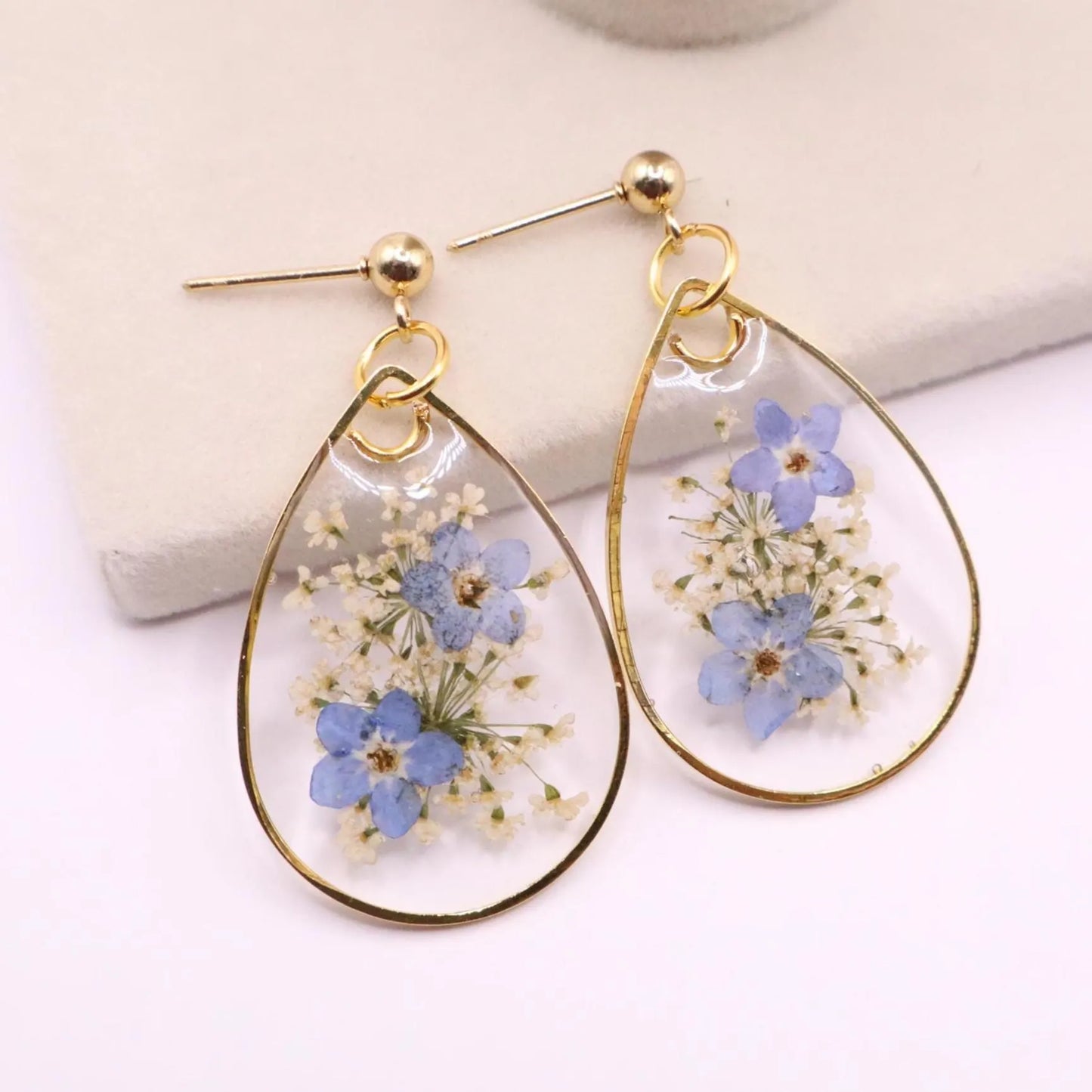 Detailed view of resin earrings with real blue flowers and white filler blooms in clear teardrop design with gold trim