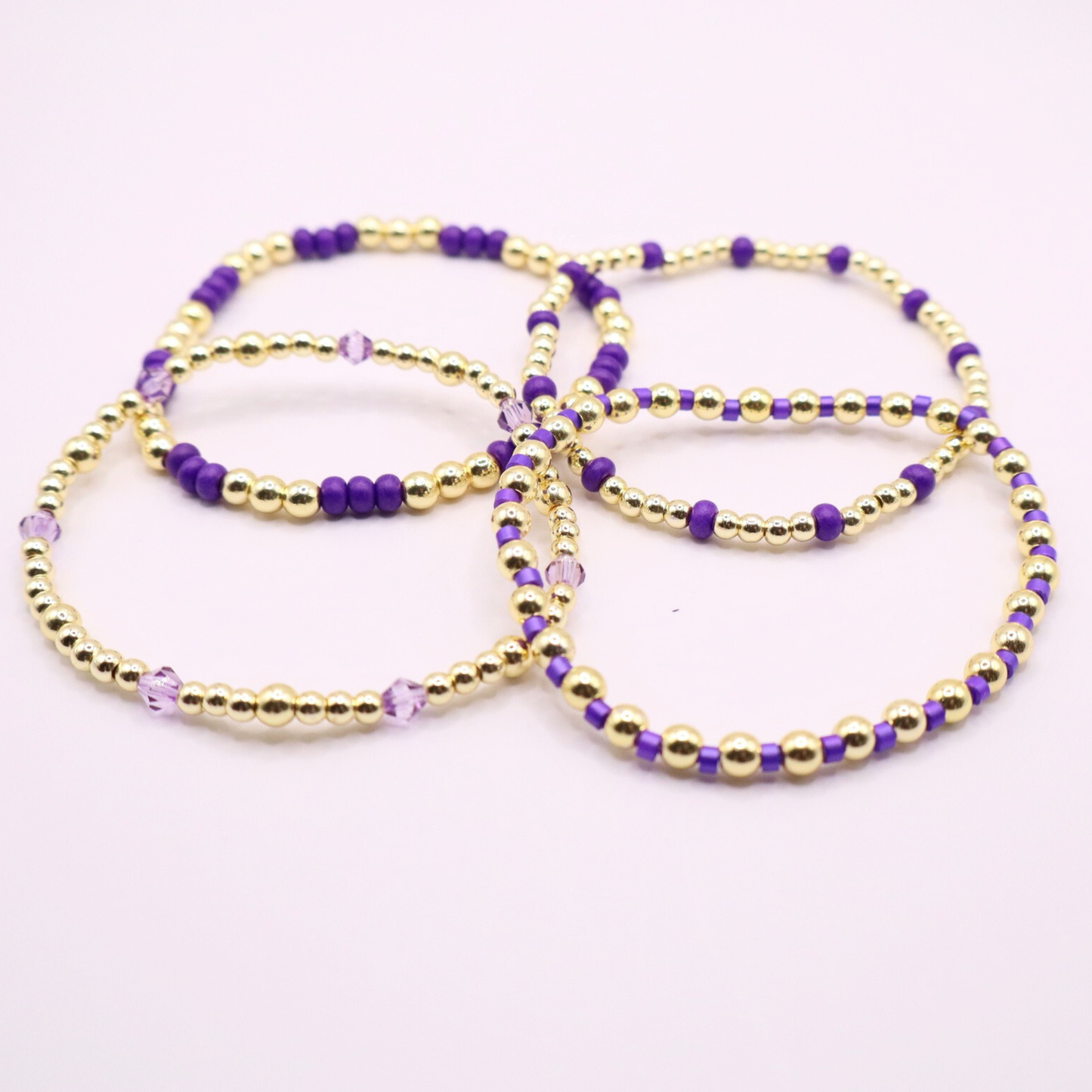 Elegant stack of stretch bracelets featuring rich purple Preciosa seed beads, faceted crystal details, and gold-tone hematite accent beads on a clean white background.