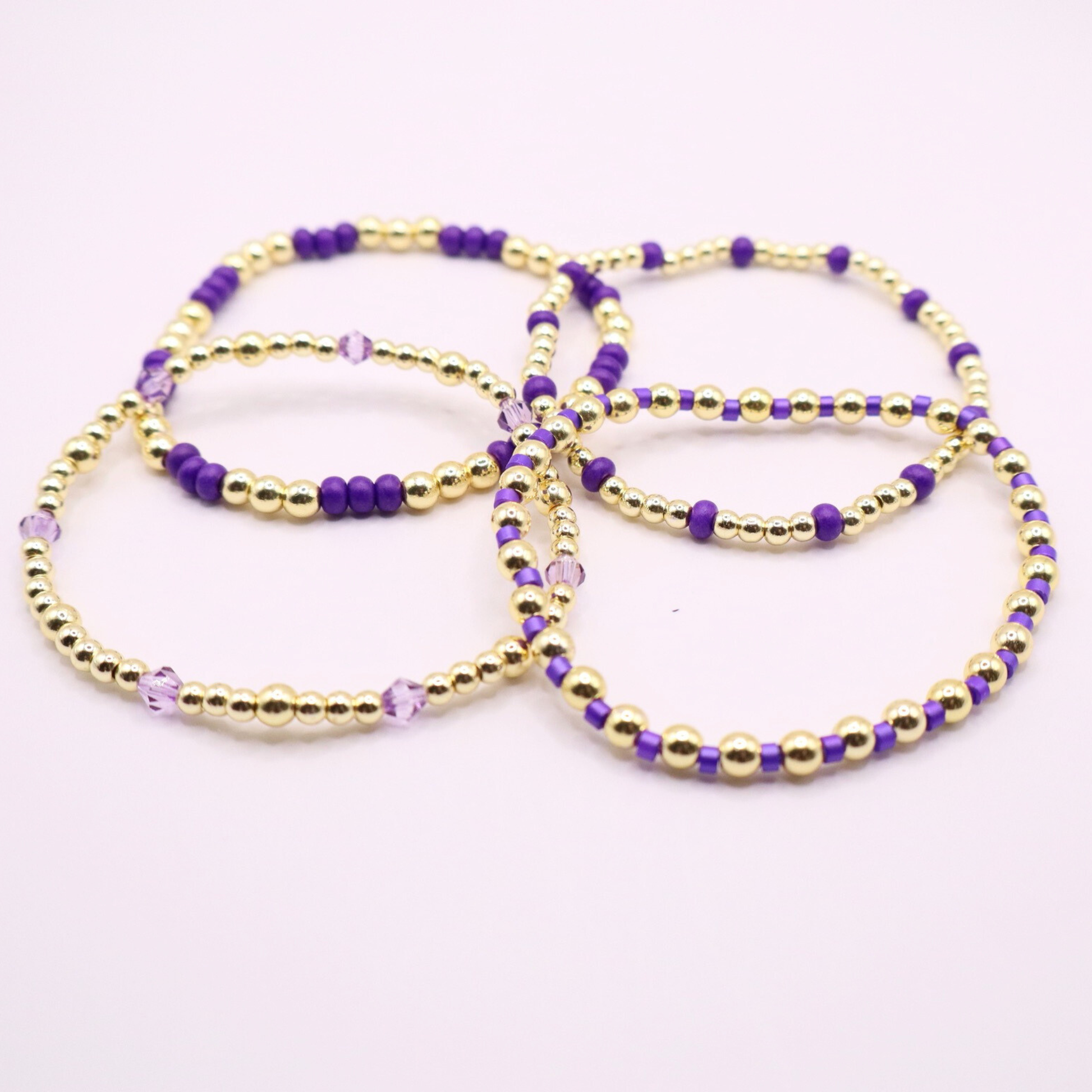 Elegant stack of stretch bracelets featuring rich purple Preciosa seed beads, faceted crystal details, and gold-tone hematite accent beads on a clean white background.