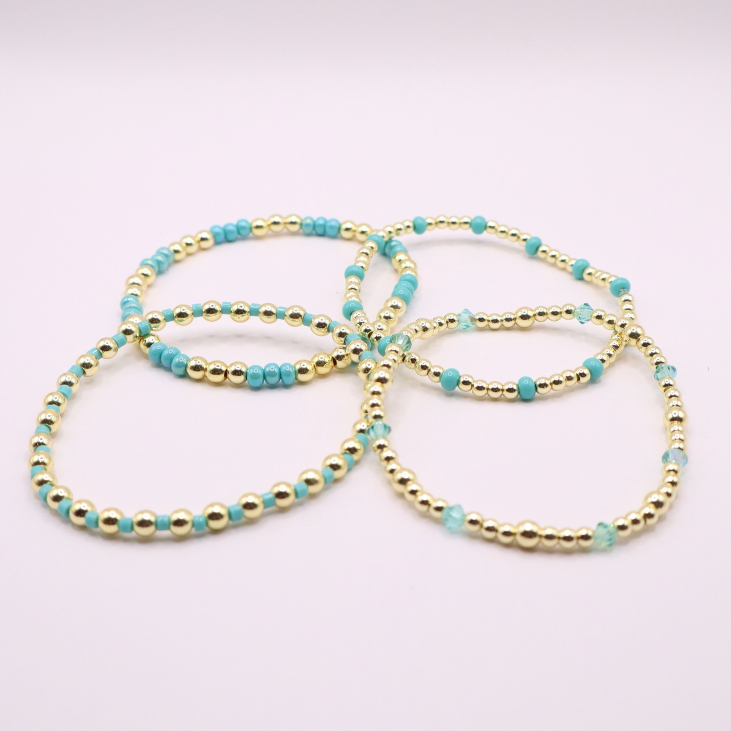 Layered bracelet stack with turquoise Preciosa seed beads, shimmering crystal accents, and gold-tone hematite beads on a clean white background.