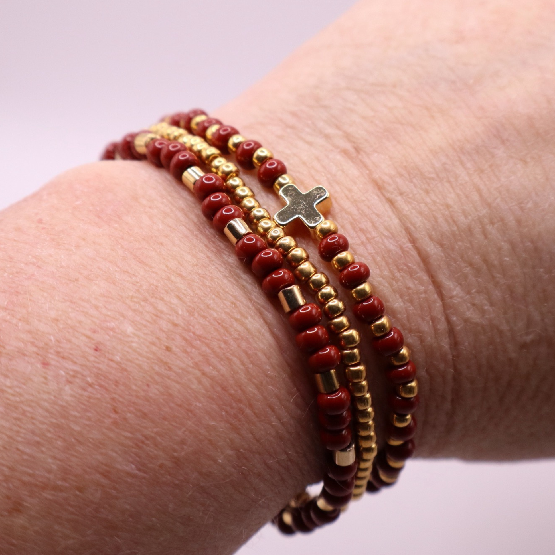 Close-up of a wrist wearing three rust and gold colored beaded bracelets. One with a cross charm on a light background