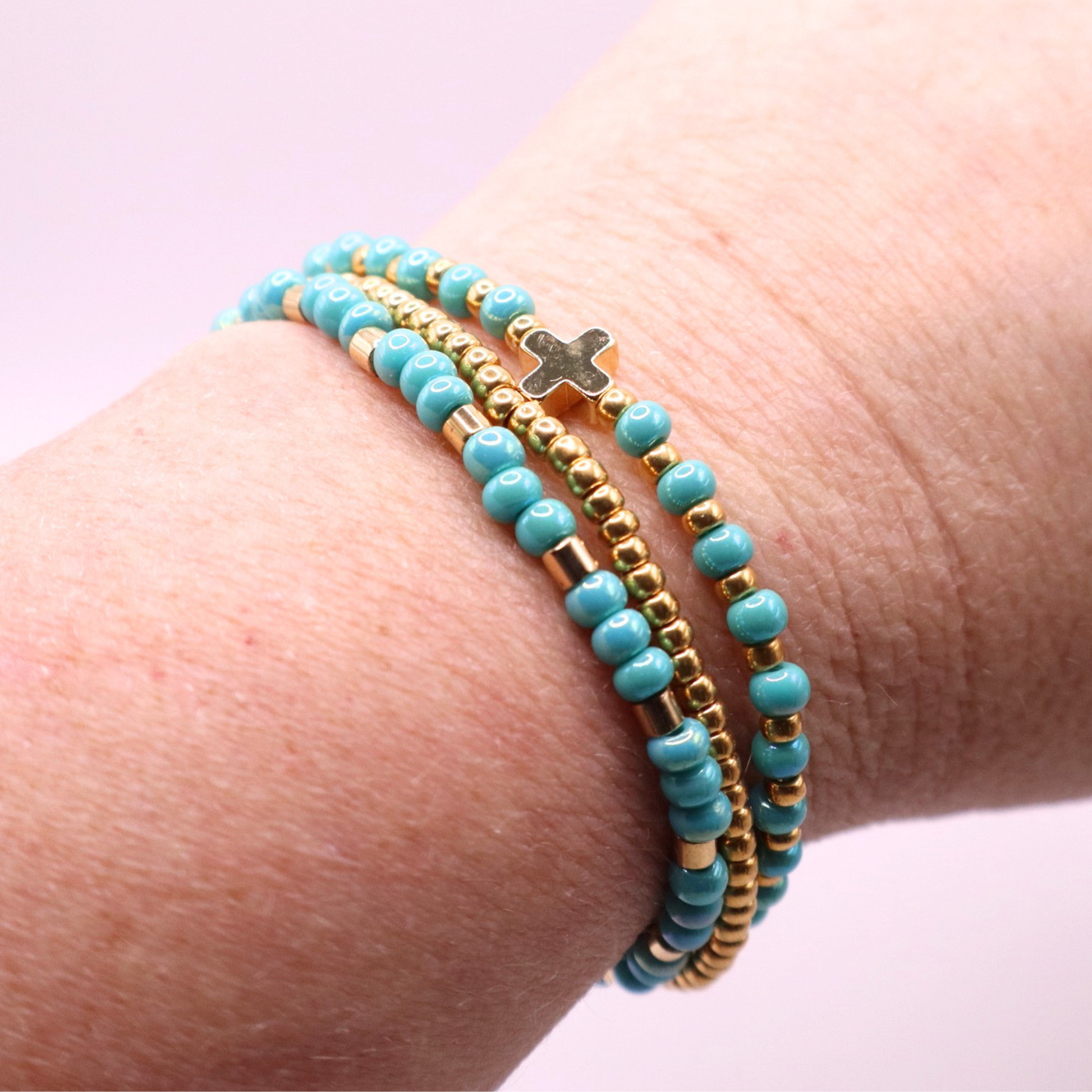 Turquoise beaded bracelet stack with gold accents and a gold cross charm on a wrist against a light pink background