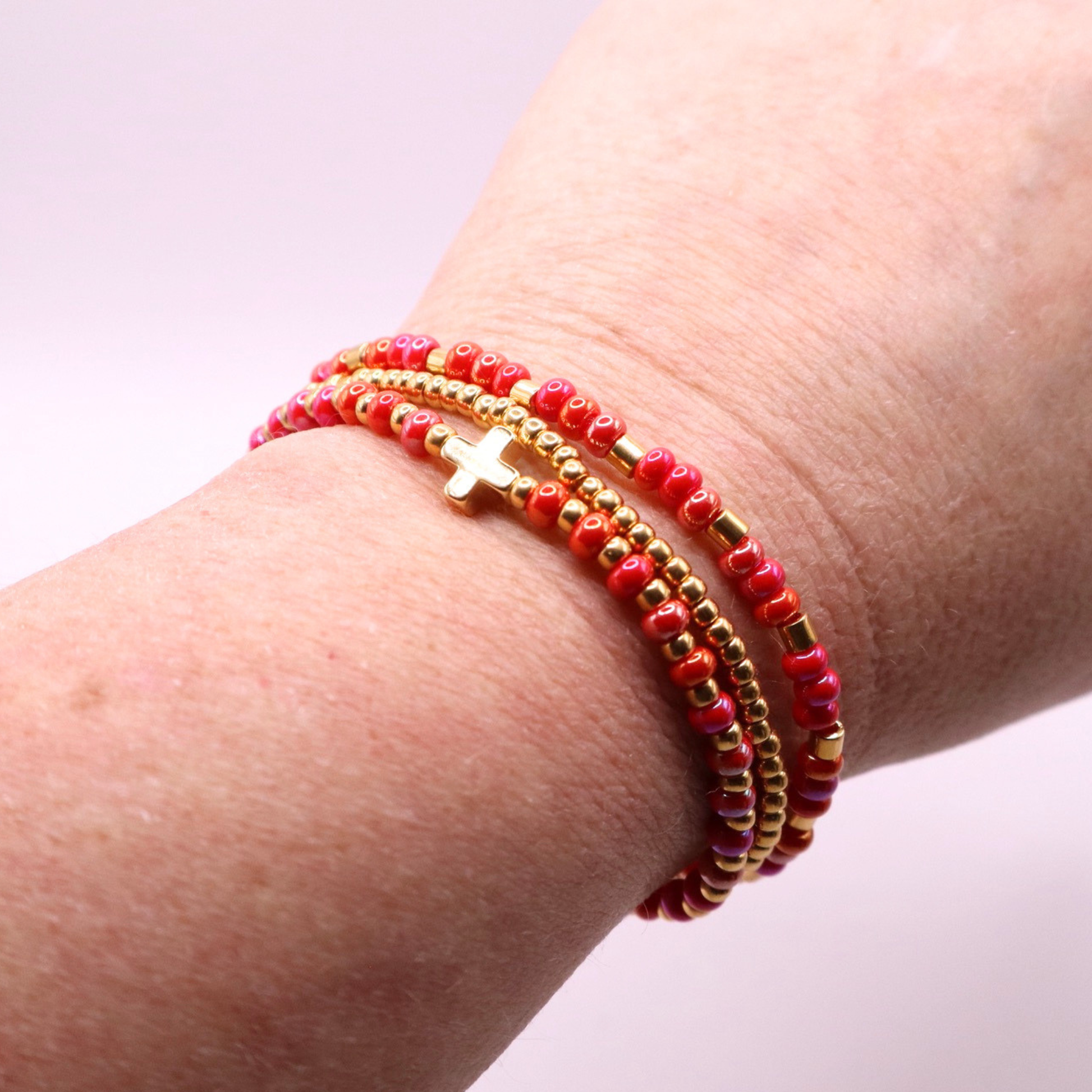 Close-up of a wrist wearing three pink and gold beaded bracelets with a cross charm on a light background