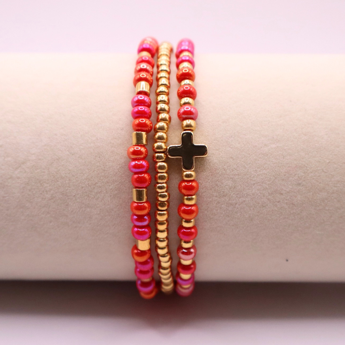 Three beaded bracelets with pink and gold beads and a gold cross charm on a beige surface.