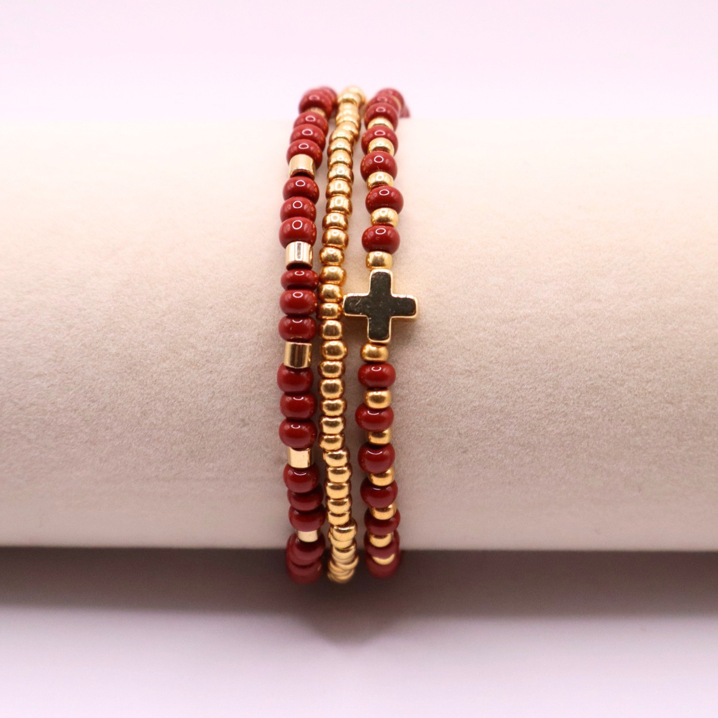 Three beaded bracelets with rust and gold beads and a gold cross charm on a beige surface