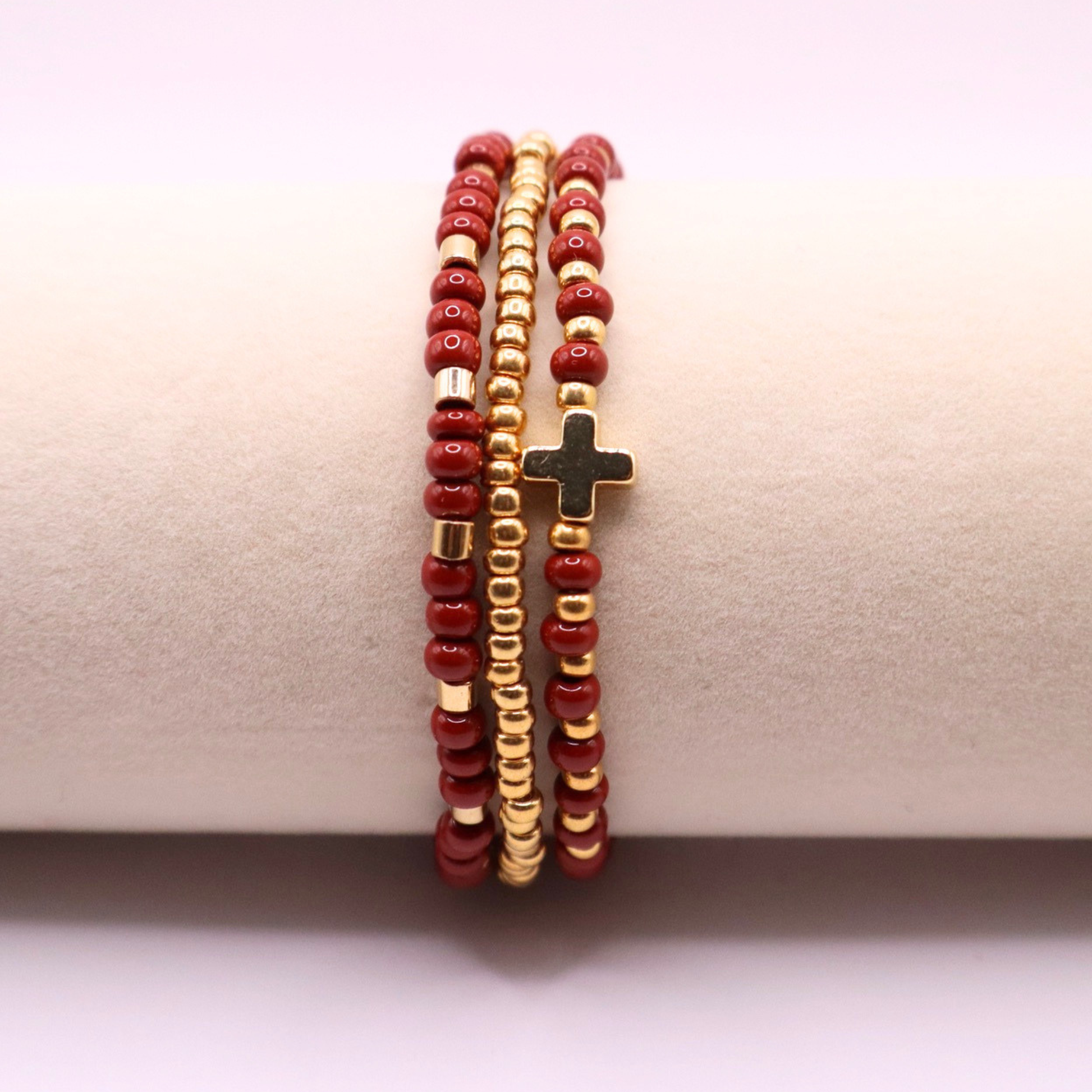 Three beaded bracelets with rust and gold beads and a gold cross charm on a beige surface