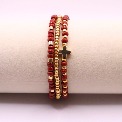 Three beaded bracelets with rust and gold beads and a gold cross charm on a beige surface