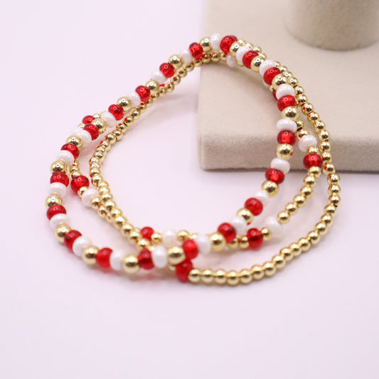 three beaded bracelets featuring various patterns of red, white and gold beads on a light pink background.
