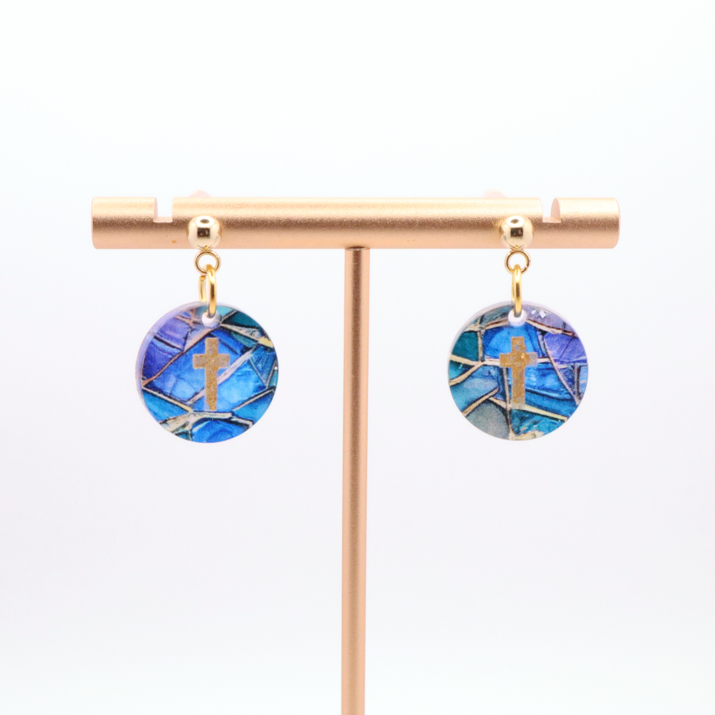 Faith inspired acrylic earrings with gold cross on blue patterned discs hanging from stainless steel stud posts on display stand