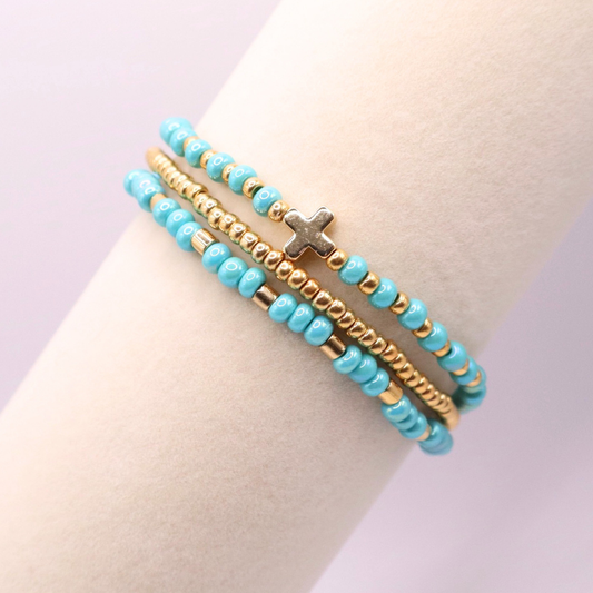 Turquoise beaded bracelet with gold accents and a cross charm on a bracelet stand against a light background