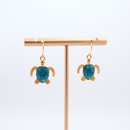Tortoise-shaped earrings with turquoise resin on a gold earring stand against a white background