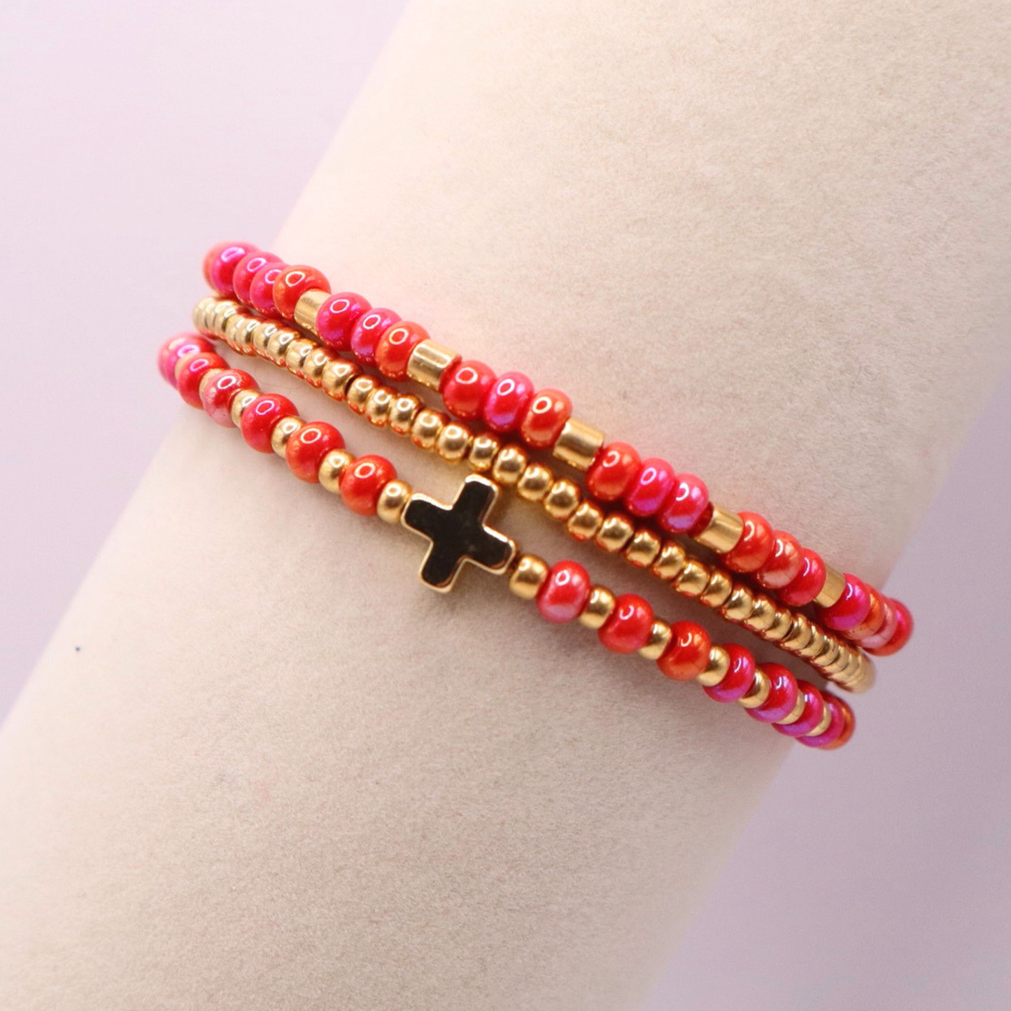 Three beaded bracelets with gold and pink beads and a gold cross charm on a mannequin arm against a light purple background.