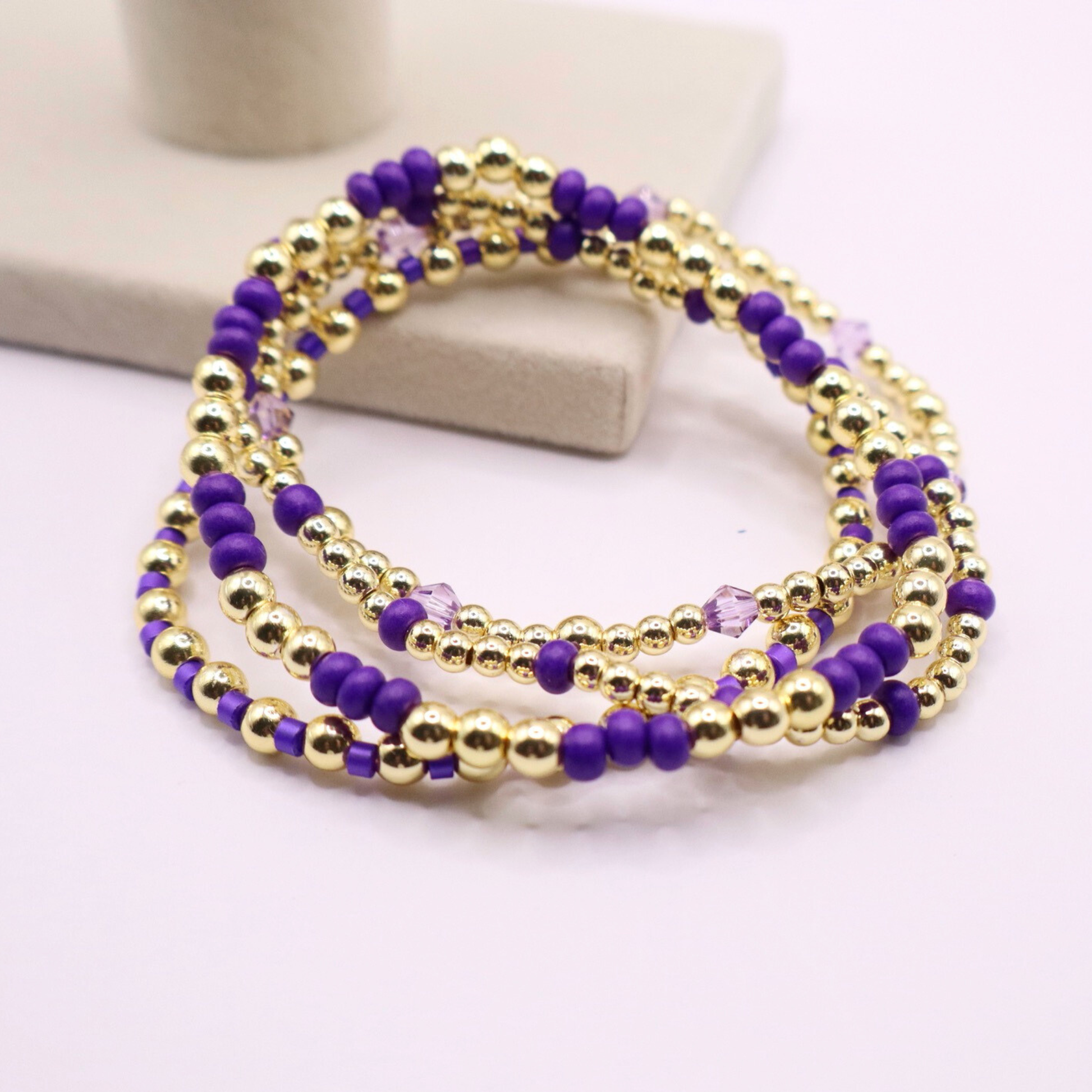 Elegant stack of stretch bracelets featuring rich purple Preciosa seed beads, faceted crystal details, and gold-tone hematite accent beads on a clean white background.