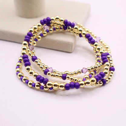 Elegant stack of stretch bracelets featuring rich purple Preciosa seed beads, faceted crystal details, and gold-tone hematite accent beads on a clean white background.