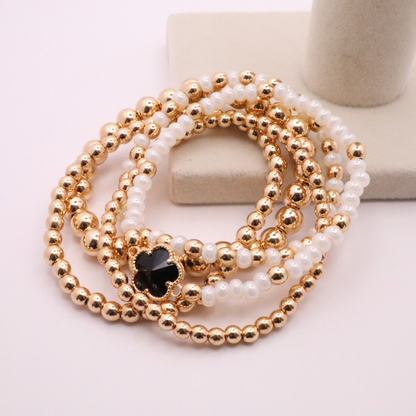 Set of 5 of gold toned beaded bracelets with white seed beads and hematite gold accents. One bracelet features a black clover charm. The bracelets are displayed on a white background.