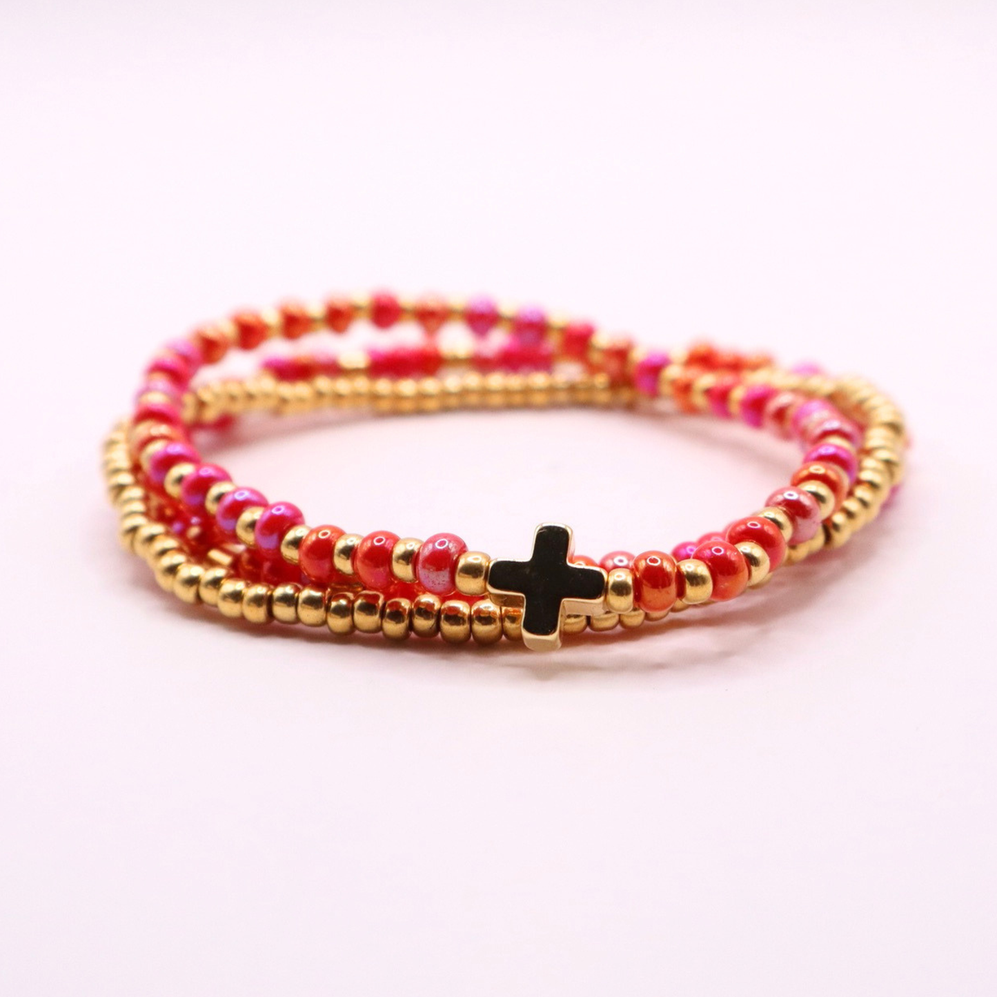 Pink beaded bracelet stack with gold accents and a cross charm on a light pink background