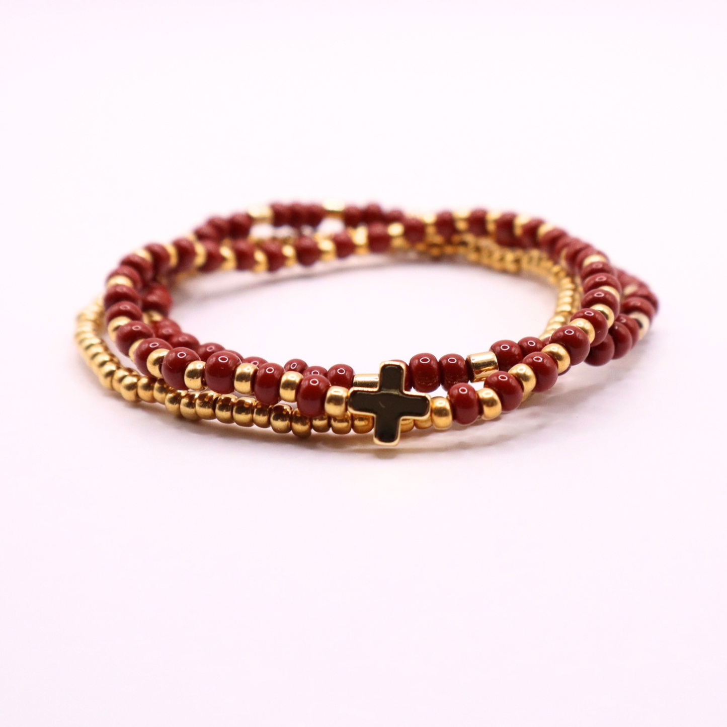 Beaded bracelet stack with gold and rust beads and a cross charm on a white background