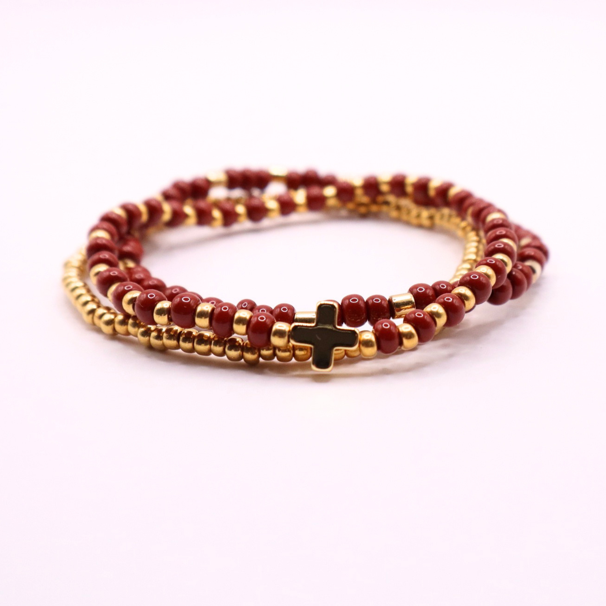 Beaded bracelet stack with gold and rust beads and a cross charm on a white background