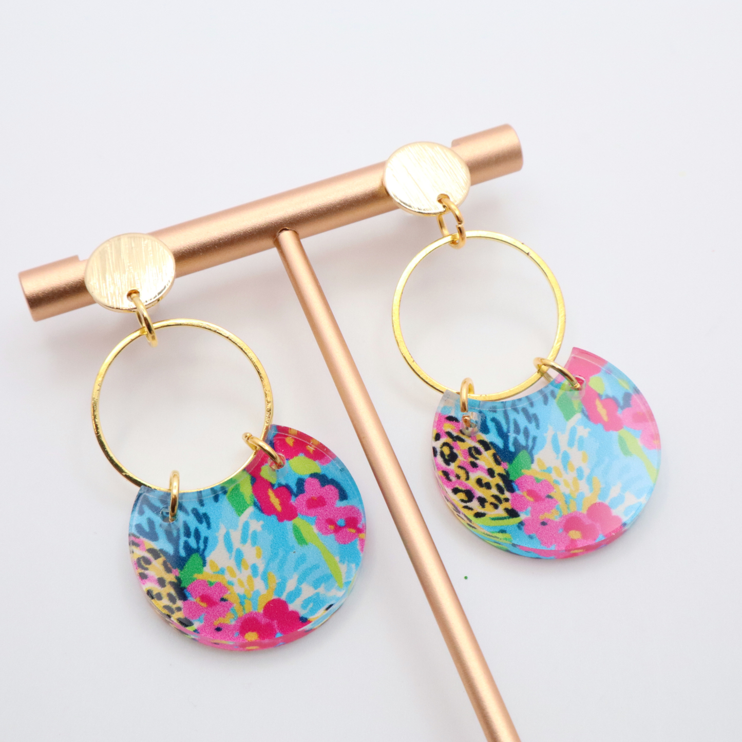 Vibrant patterned acrylic earrings with multicolor floral design hanging from gold hoops and 18k gold plated posts on a jewelry stand