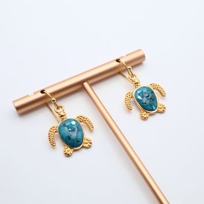 Turtle-shaped earrings with teal blue resin on gold hooks, displayed on a white background.