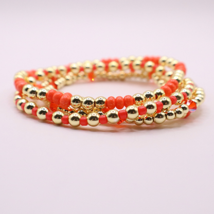 Stack of handmade stretch bracelets featuring vibrant coral Preciosa seed beads, faceted crystal accents, and polished gold-tone hematite beads on a clean white background.