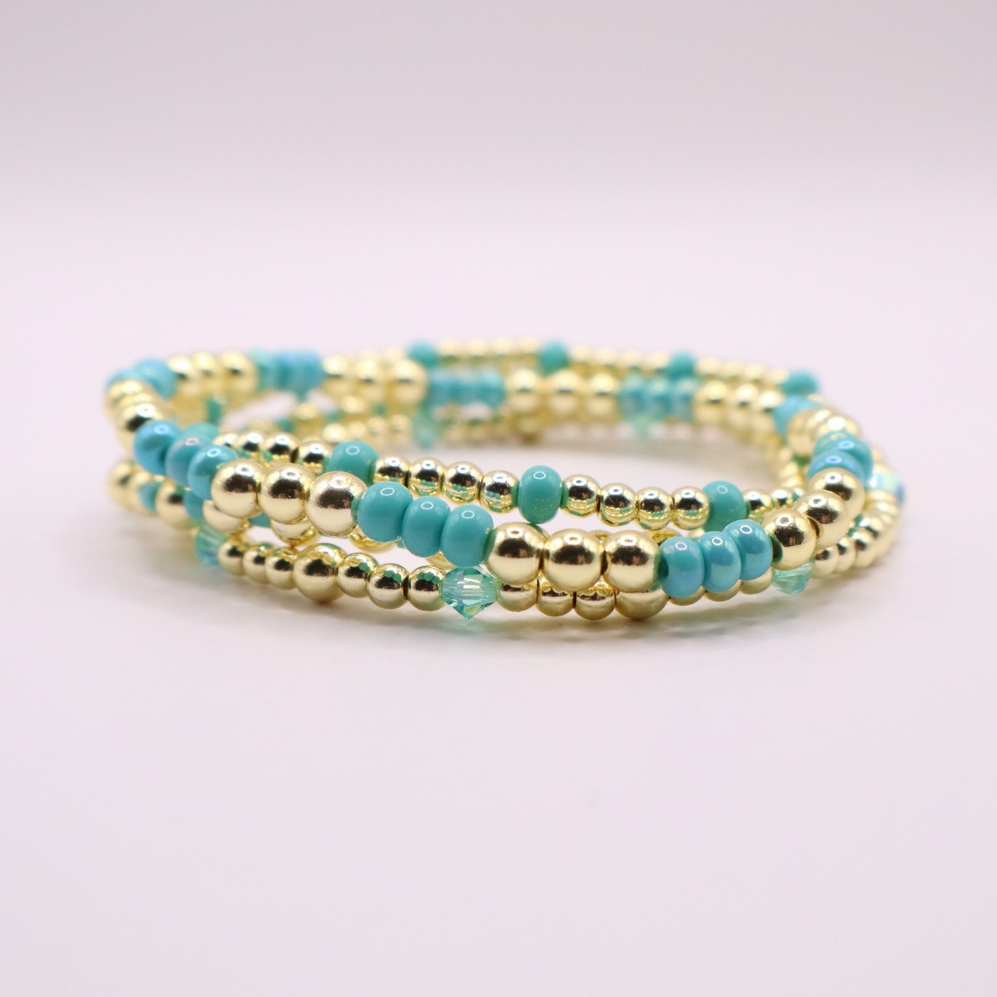 Layered bracelet stack with turquoise Preciosa seed beads, shimmering crystal accents, and gold-tone hematite beads on a clean white background.