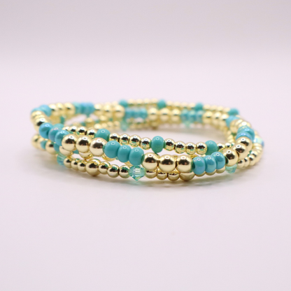 Layered bracelet stack with turquoise Preciosa seed beads, shimmering crystal accents, and gold-tone hematite beads on a clean white background.