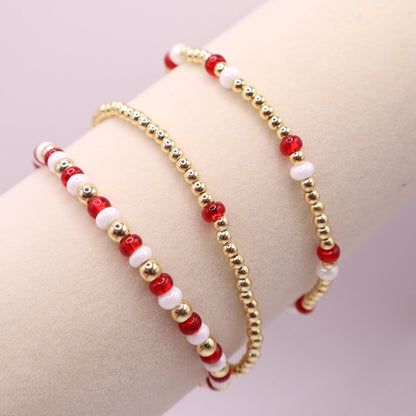 Three beaded bracelets with red, white, and gold accents on a bracelet display against a light background.
