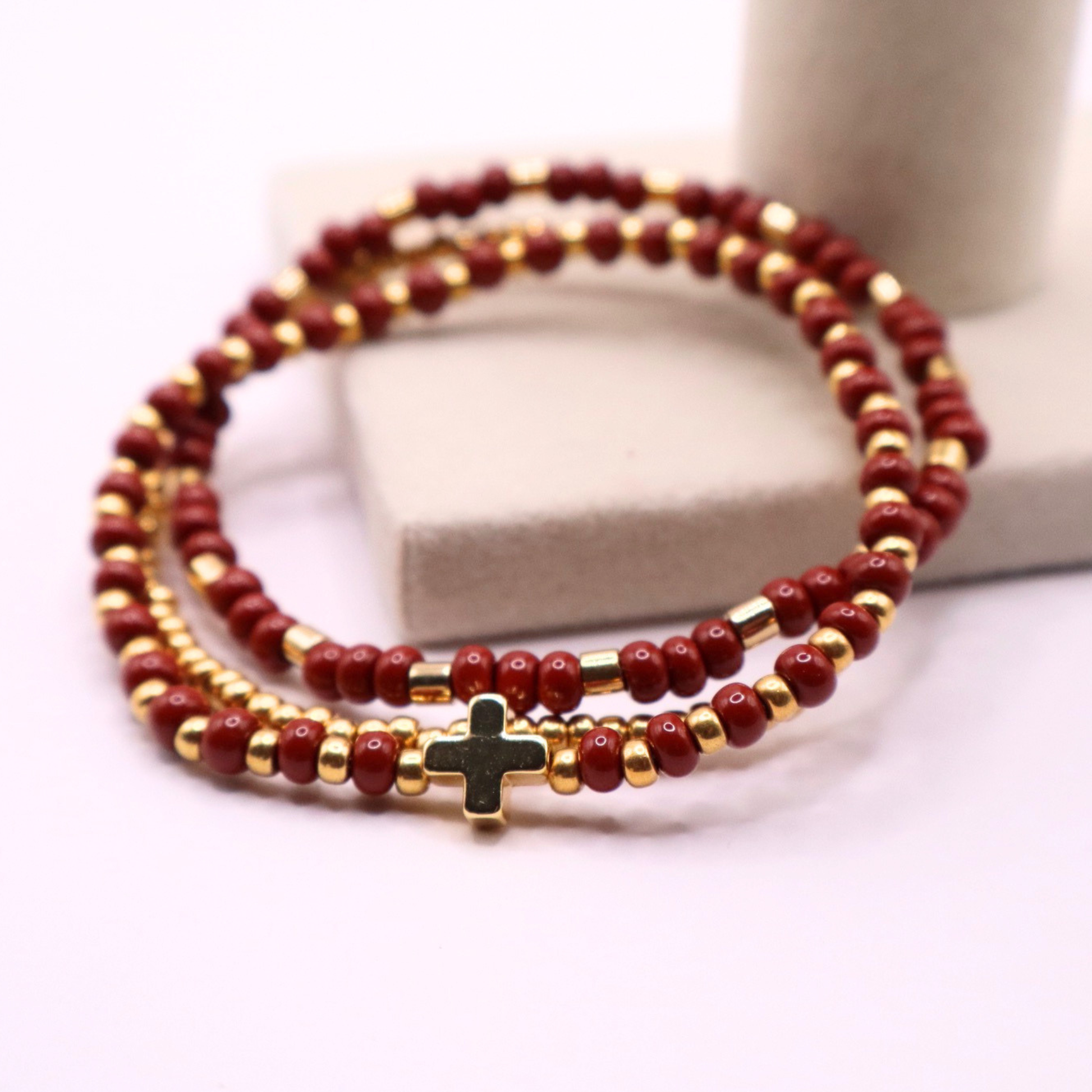 Beaded bracelet stack with rust and gold beads and a cross charm on a white background