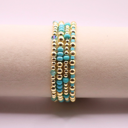 Layered bracelet stack with turquoise Preciosa seed beads, shimmering crystal accents, and gold-tone hematite beads on a clean white background.