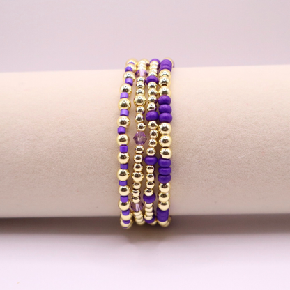 Elegant stack of stretch bracelets featuring rich purple Preciosa seed beads, faceted crystal details, and gold-tone hematite accent beads on a clean white background.