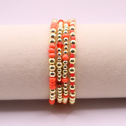 Stack of handmade stretch bracelets featuring vibrant coral Preciosa seed beads, faceted crystal accents, and polished gold-tone hematite beads on a clean white background.