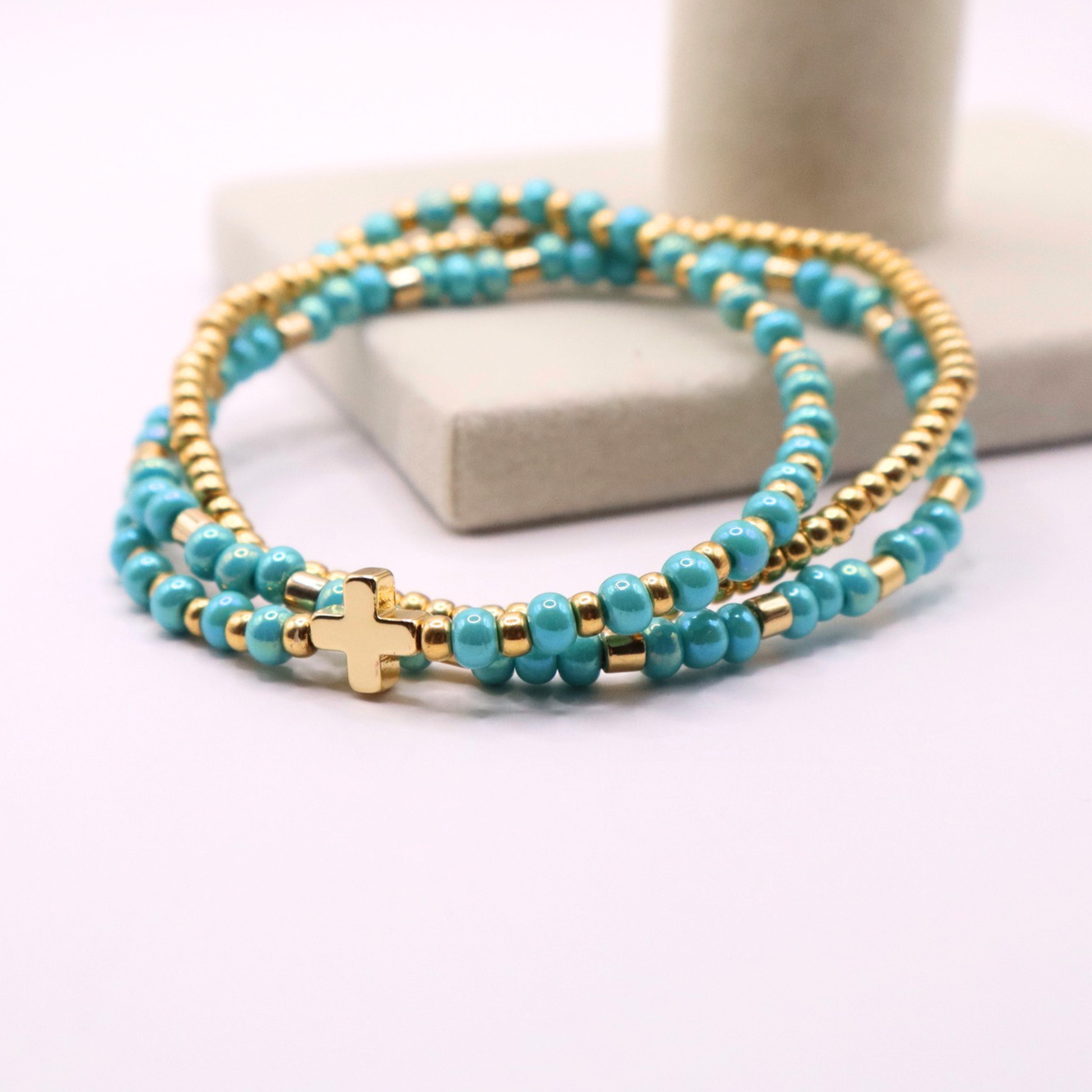 Turquoise beaded bracelet stack with gold accents and a gold cross charm on a white surface