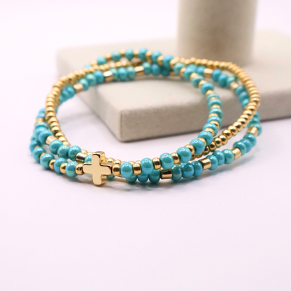 Turquoise beaded bracelet stack with gold accents and a gold cross charm on a white surface