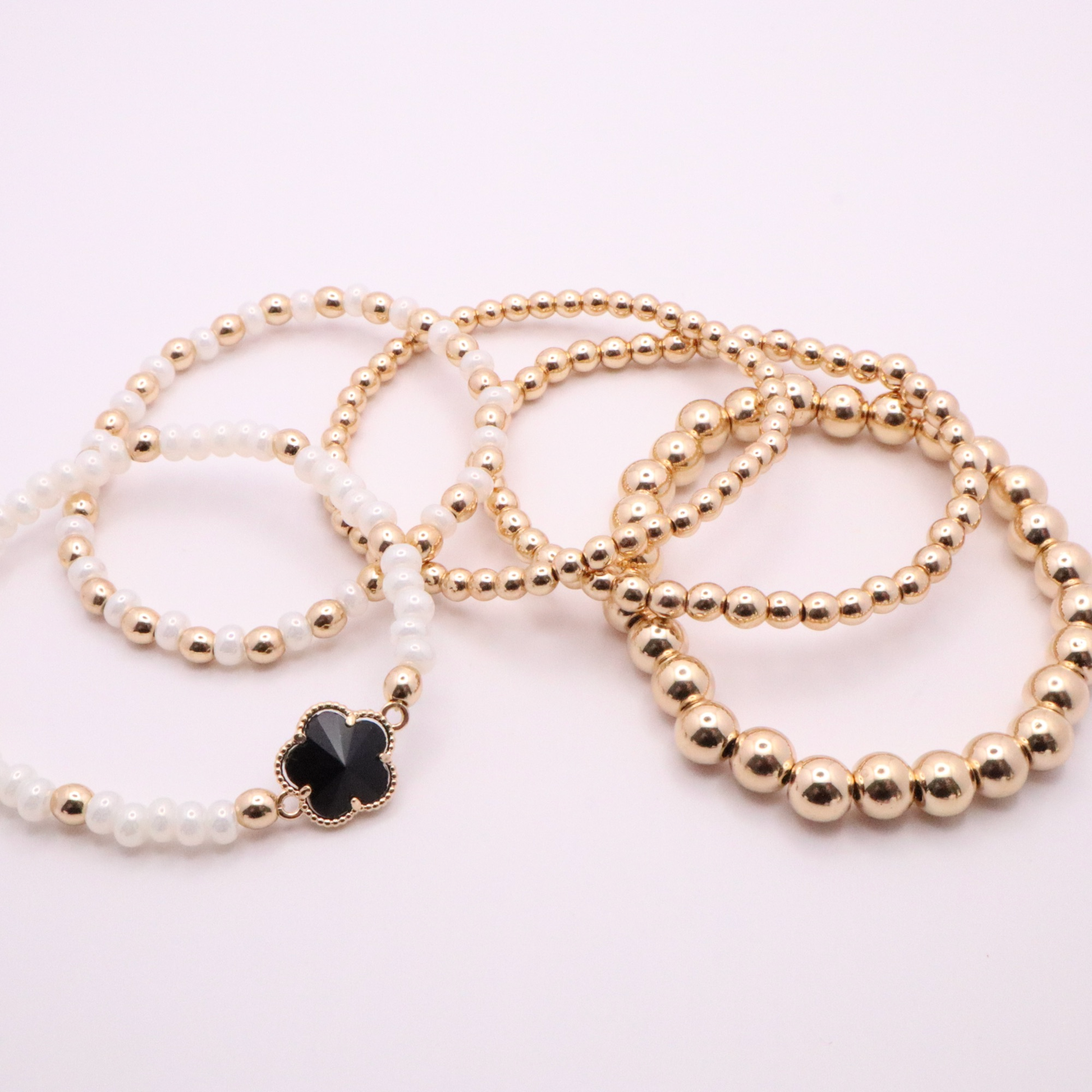 Set of 5 of gold toned beaded bracelets with white seed beads and hematite gold accents. One bracelet features a black clover charm. The bracelets are displayed on a white background.