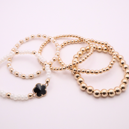 Set of 5 of gold toned beaded bracelets with white seed beads and hematite gold accents. One bracelet features a black clover charm. The bracelets are displayed on a white background.