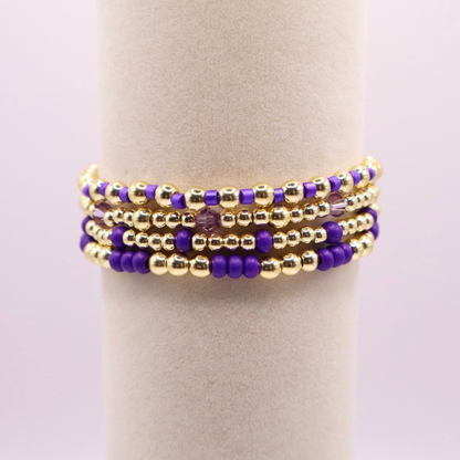 Elegant stack of stretch bracelets featuring rich purple Preciosa seed beads, faceted crystal details, and gold-tone hematite accent beads on a clean white background.