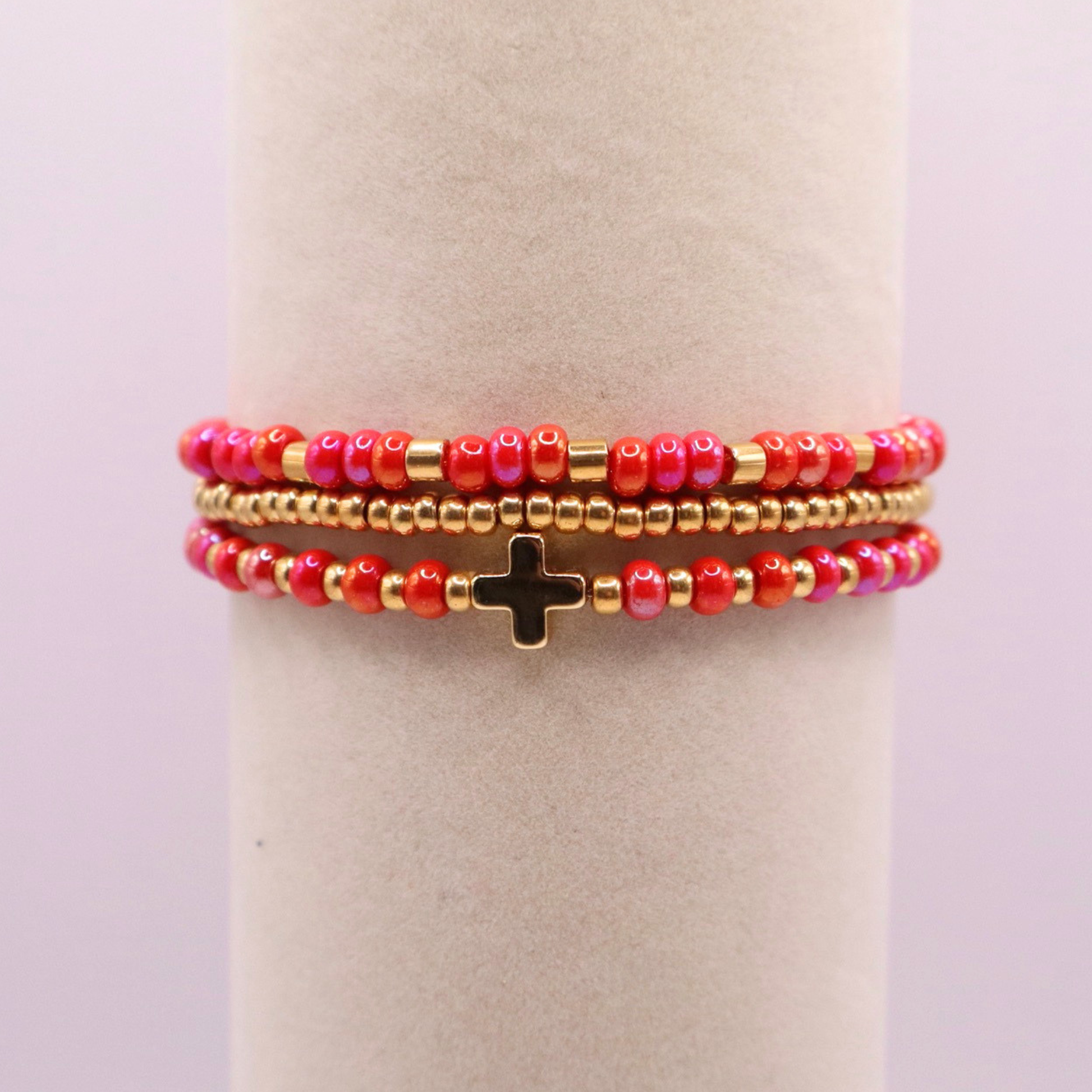 Stack of pink and gold beaded bracelets with a gold cross charm on a white cylindrical stand with a light purple background