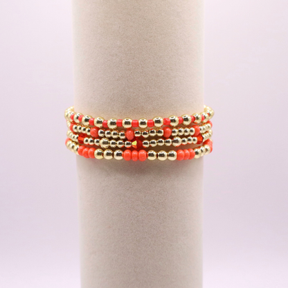 Stack of handmade stretch bracelets featuring vibrant coral Preciosa seed beads, faceted crystal accents, and polished gold-tone hematite beads on a clean white background.