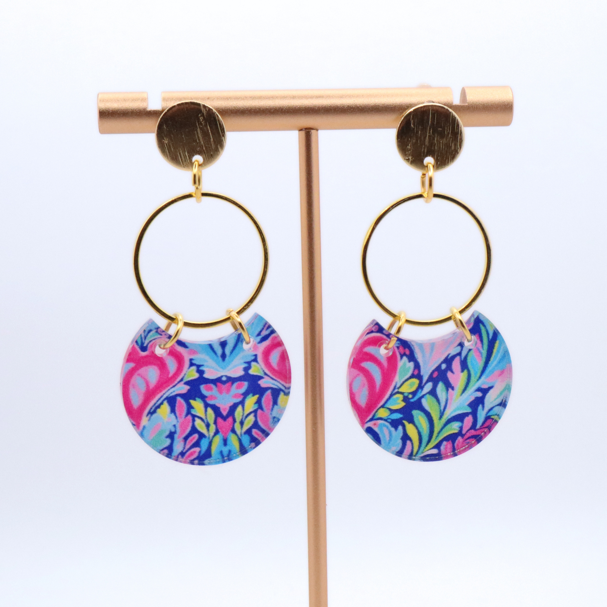 Bright patterned acrylic earrings with pink, blue, and green design on gold hoops with 18k gold plated posts displayed on stand