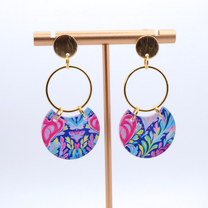 Bright patterned acrylic earrings with pink, blue, and green design on gold hoops with 18k gold plated posts displayed on stand
