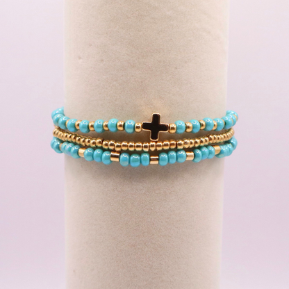 Turquoise beaded bracelet stack with gold accents and a cross charm on a white stand.