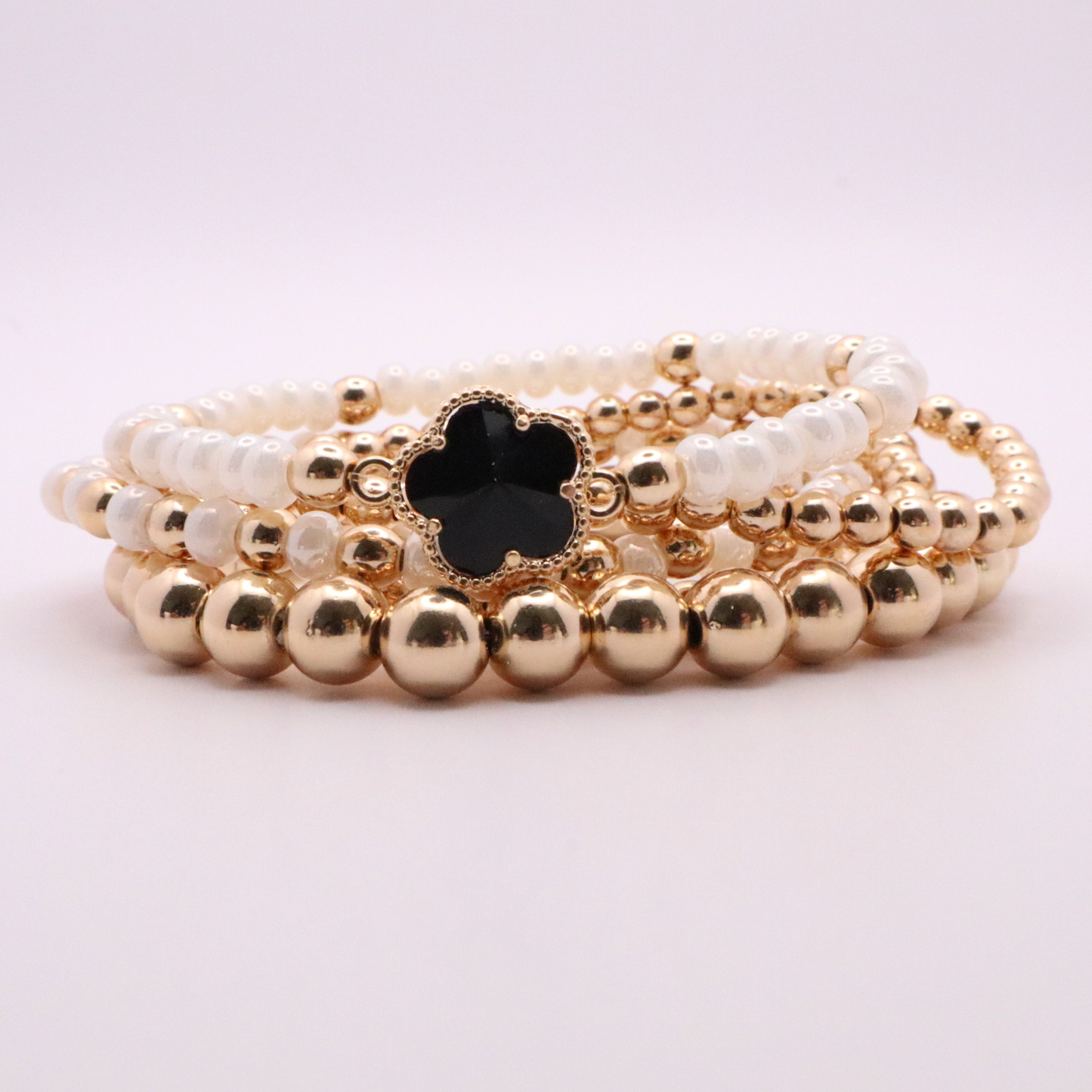 Set of 5 of gold toned beaded bracelets with white seed beads and hematite gold accents. One bracelet features a black clover charm. The bracelets are displayed on a white background.