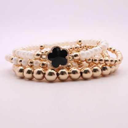 Set of 5 of gold toned beaded bracelets with white seed beads and hematite gold accents. One bracelet features a black clover charm. The bracelets are displayed on a white background.