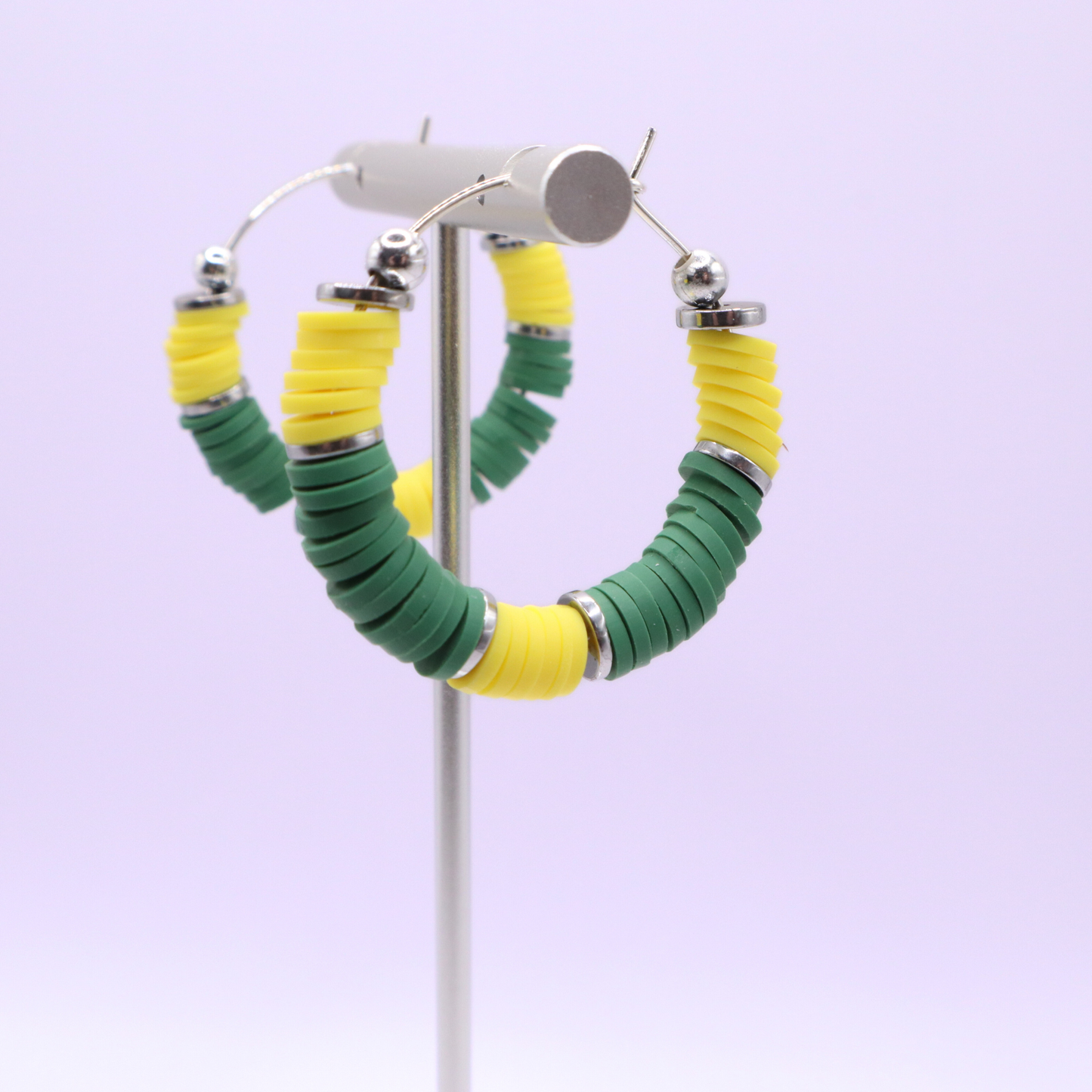 Green and yellow heishi bead hoop earrings with silver accents hanging on a minimalist jewelry stand against a white background
