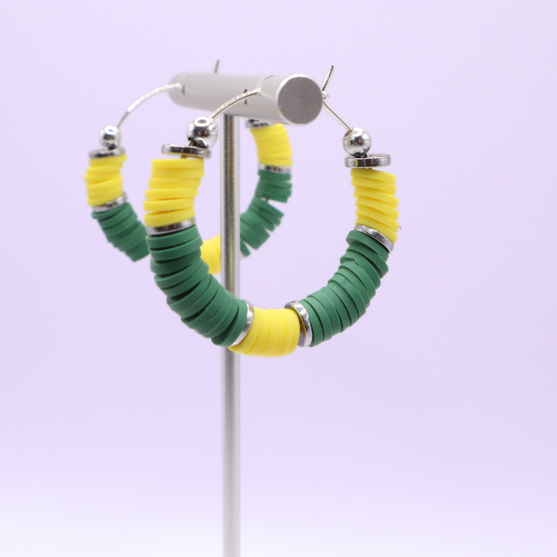 Green and yellow heishi bead hoop earrings with silver accents hanging on a minimalist jewelry stand against a white background