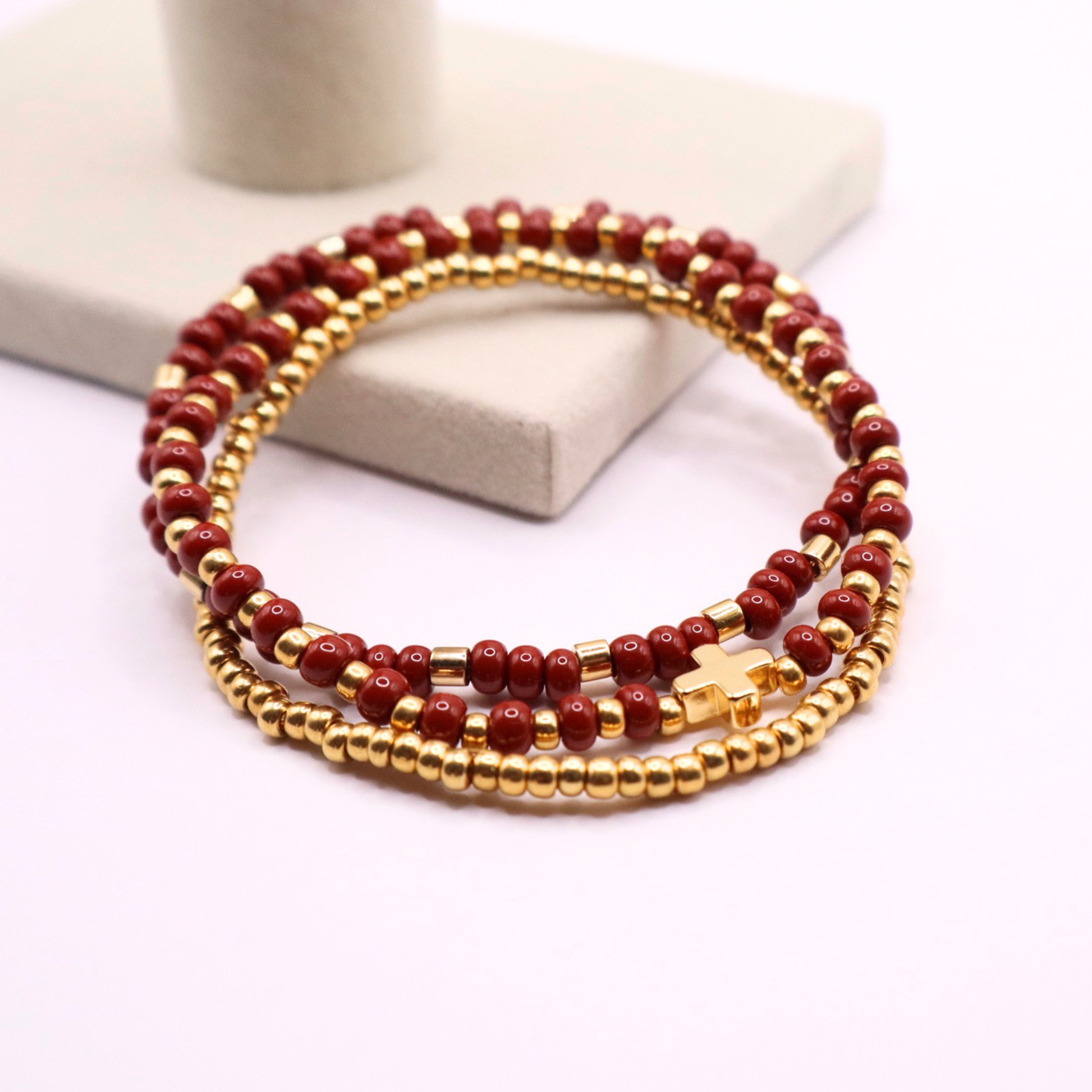 Stack of rust and gold beaded bracelets, one with a gold gross charm, on a white surface